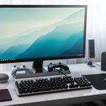 Modern desktop setup with large monitor, keyboard, mouse, game controllers, speakers, and a potted plant on a white desk